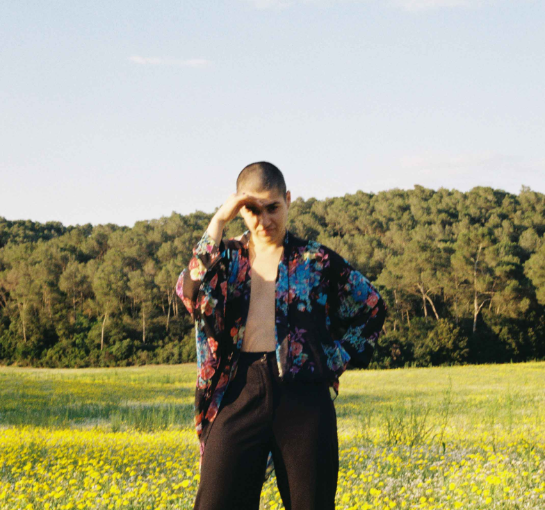 El artista de rap Xeno Bitch aparece de pie en el bosque mirando de frente, tapándose los ojos del sol con la mano